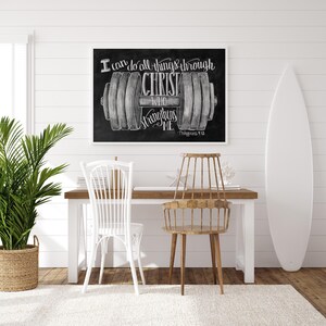 May include: A framed chalkboard art print with the text "I can do all things through Christ who strengthens me" and a dumbbell illustration. The print is displayed above a wooden desk with two chairs and a surfboard.