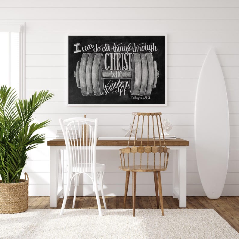 May include: A framed chalkboard art print with the text "I can do all things through Christ who strengthens me" and a dumbbell illustration. The print is displayed above a wooden desk with two chairs and a surfboard.
