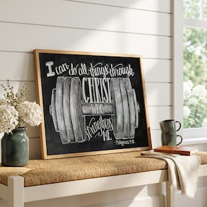May include: A framed chalkboard with a wooden frame displays the text "I can do all things through Christ who strengthens me." A dumbbell graphic is centered below the text. A vase of white flowers sits to the left.