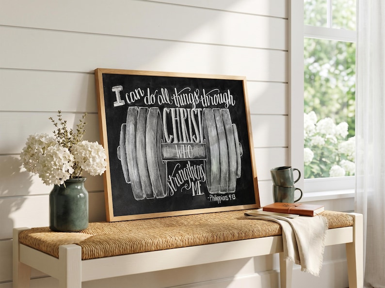 May include: A framed chalkboard with a wooden frame displays the text "I can do all things through Christ who strengthens me." A dumbbell graphic is centered below the text. A vase of white flowers sits to the left.