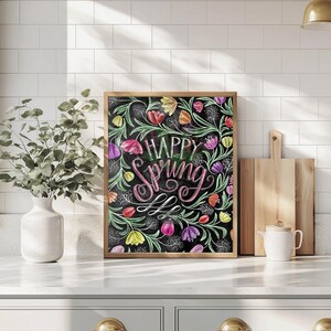 May include: A framed artwork with the words "Happy Spring" in a decorative script. The image shows colorful tulips and floral elements on a black background, within a wooden frame. The artwork is displayed on a white countertop.