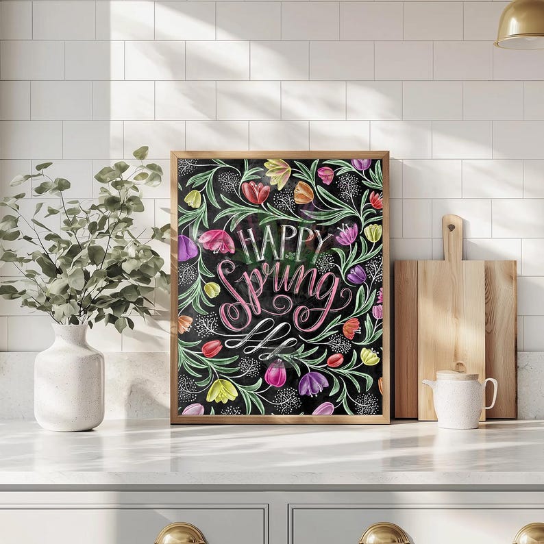 May include: A framed artwork with the words "Happy Spring" in a decorative script. The image shows colorful tulips and floral elements on a black background, within a wooden frame. The artwork is displayed on a white countertop.