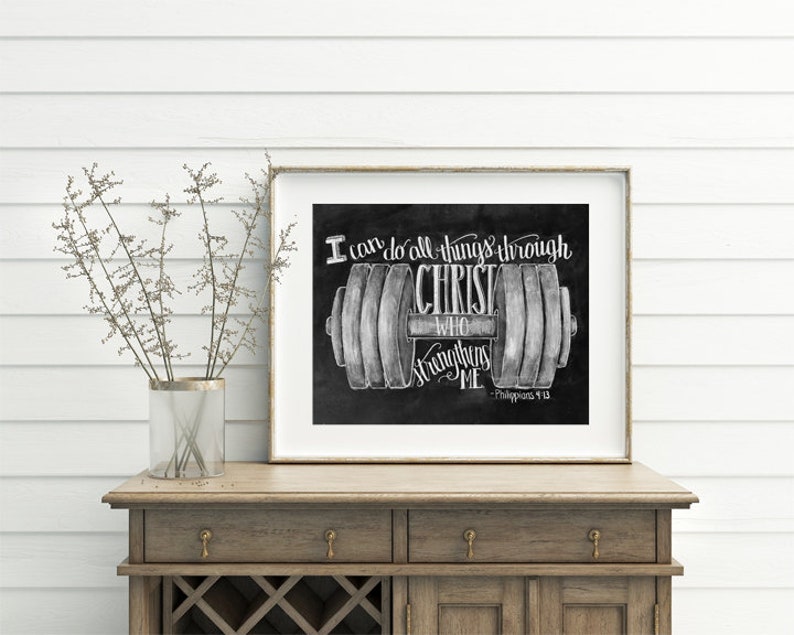 May include: Framed black and white print featuring a barbell design with the text "I can do all things through Christ who strengthens me." The print is displayed on a wooden cabinet with a vase of dried flowers.