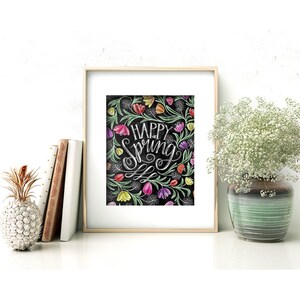 May include: A framed art print with the words "Happy Spring" in white script, surrounded by colourful flowers and green leaves on a black background. The print is in a gold frame and is displayed on a white surface with books and a decorative pineapple.