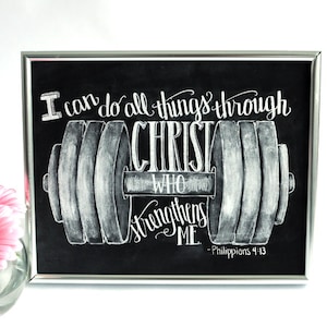 May include: Black framed chalkboard art with white lettering. The text reads "I can do all things through Christ who strengthens me - Philippians 4:13" with a barbell illustration. A pink flower is in the background.