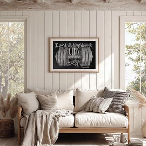 May include: A framed print with the text "I can do all things through Christ who strengthens me" and a barbell illustration. The print is displayed above a light-colored wooden sofa with neutral-toned pillows and a throw blanket.