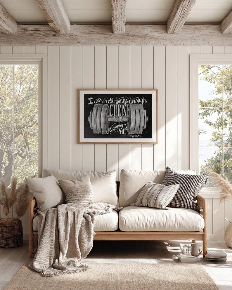 May include: A framed print with the text "I can do all things through Christ who strengthens me" and a barbell illustration. The print is displayed above a light-colored wooden sofa with neutral-toned pillows and a throw blanket.
