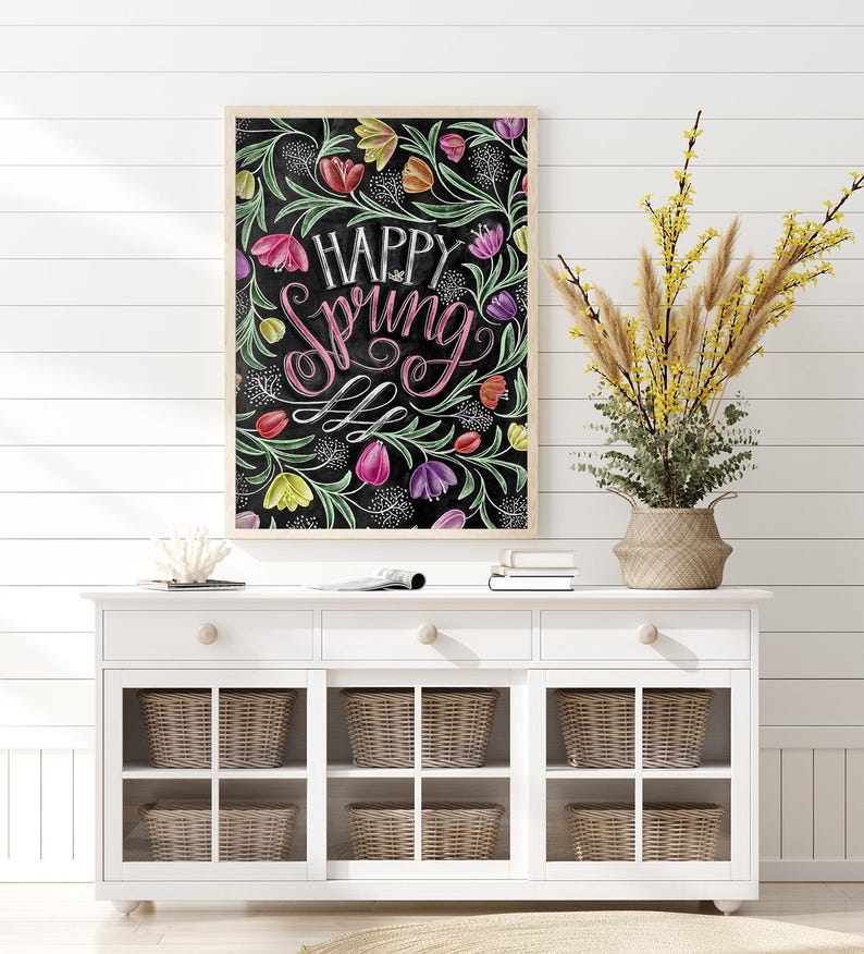 May include: A framed art print with the words "Happy Spring" in white and pink script, surrounded by colourful tulips and green foliage on a black background. The print is displayed above a white cabinet with woven baskets.