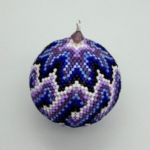 Puede incluir: Un adorno navideño de cuentas de color morado, azul, blanco y negro con un patrón geométrico. El adorno tiene un lazo plateado y una cuenta morada en la parte superior.