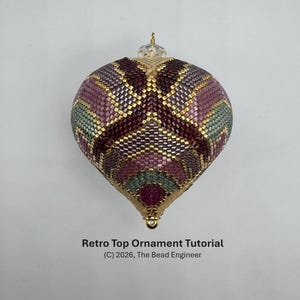 Puede incluir: Un adorno de cuentas en forma de lágrima con un patrón geométrico en tonos púrpura oscuro, malva y verde salvia con detalles dorados. Una cuenta de cristal transparente se sitúa en la parte superior. Texto: Retro Top Ornament Tutorial (C) 2026, The Bead Engineer.