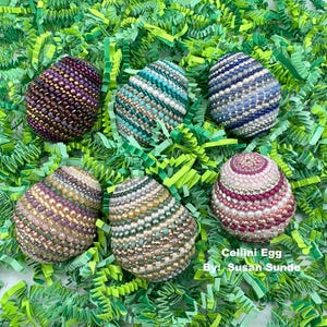 May include: Six intricately beaded Easter eggs in various colors, including purple, blue, green, pink, and gold. The eggs are arranged on a bed of green shredded paper. The text "Cellini Egg By: Susan Sunde" is visible at the bottom of the image.