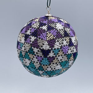 Puede incluir: Un adorno de Navidad de cuentas con un patrón geométrico en tonos de morado, azul verdoso, blanco y plateado.