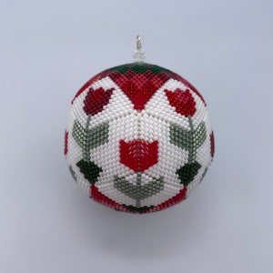 Puede incluir: Un adorno navideño de cuentas blancas, rojas y verdes con un diseño floral. El adorno es redondo y tiene un lazo en la parte superior para colgar.