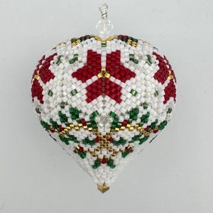 May include: A white, red, green and gold beaded Christmas bauble in the shape of a heart. The bauble has a geometric pattern with a star-like design.