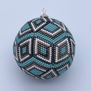 Puede incluir: Una bola ornamental con cuentas con un patrón geométrico en turquesa, blanco, gris y negro. El adorno tiene un lazo de metal plateado en la parte superior para colgar. El trabajo de cuentas crea una superficie texturizada.
