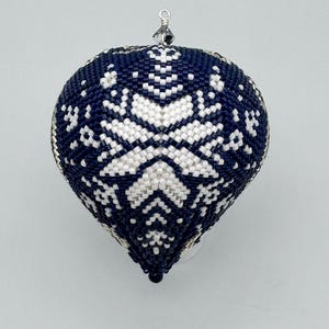 Op de afbeelding: Een marineblauwe en witte kerstversiering van kralen met een sneeuwvlokontwerp. De druppelvormige versiering heeft een zilverkleurige metalen dop met een kristallen kraal. De versiering is ongeveer 7.6 cm hoog.