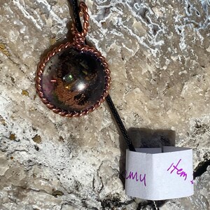 May include: A handcrafted pendant necklace featuring a round, clear glass orb with dark, speckled inclusions and iridescent accents. The orb is encased in a copper-colored, twisted wire frame and hangs from a black cord. A small tag with handwritten text is attached.