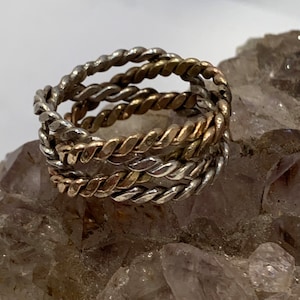 May include: A stack of three twisted metal rings in silver and gold tones. The rings are tightly coiled, creating a rope-like texture. The rings are stacked on a rough, light brown surface.