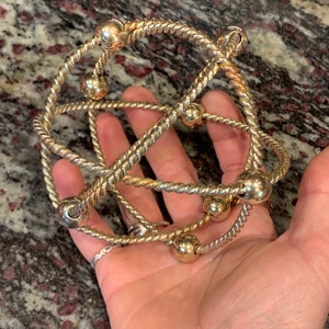 May include: A gold-toned metal bracelet with a twisted rope design and round beads. The bracelet is made of multiple strands of rope that are intertwined and connected by the beads.