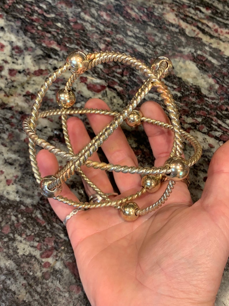 May include: A gold-toned metal bracelet with a twisted rope design and round beads. The bracelet is made of multiple strands of rope that are intertwined and connected by the beads.
