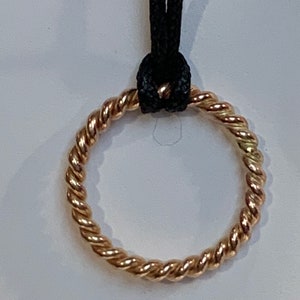 Op de afbeelding: Een goudkleurige hangerketting met een gedraaid touwontwerp. De cirkelvormige hanger is opgehangen aan een zwart koord. De hanger heeft een gevlochten uiterlijk, met een glanzende, metallic afwerking. De ketting wordt gepresenteerd tegen een effen witte achtergrond.