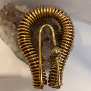 May include: A gold-colored metal brooch shaped like a horseshoe with a coiled spring design. The brooch has a small round ball at the top.
