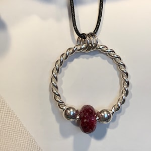 May include: A pendant necklace featuring a circular, twisted silver-tone metal ring suspended from a thin black cord. The ring is accented with a single faceted, deep red bead flanked by two smooth, polished silver-tone spherical beads.
