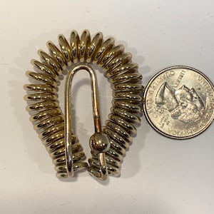 May include: A gold-colored metal key chain with a spring coil design and a hook. The key chain is next to a US quarter.