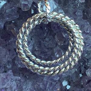 May include: A silver necklace with a twisted circle pendant. The pendant is made of three intertwined circles, creating a unique and modern design. The necklace is photographed against a background of amethyst crystals.