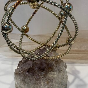 May include: A silver wire sculpture with multiple intertwined rings and silver beads, resting on a large amethyst crystal.