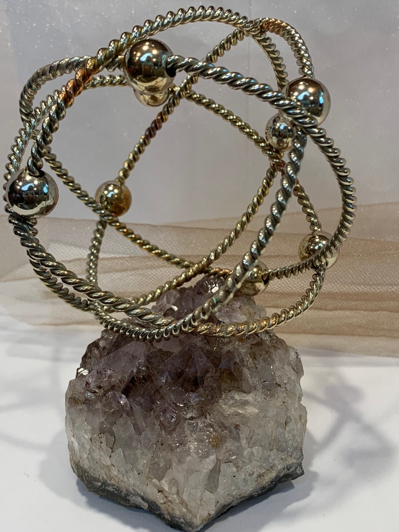 May include: A silver wire sculpture with multiple intertwined rings and silver beads, resting on a large amethyst crystal.