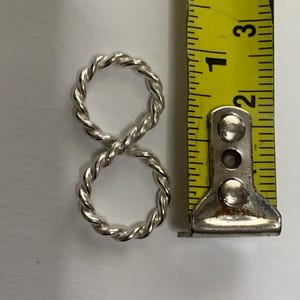 May include: A silver-colored twisted metal ring shaped like an infinity symbol, positioned next to a yellow measuring tape that shows the object is approximately 1.25 inches in total height from top to bottom.