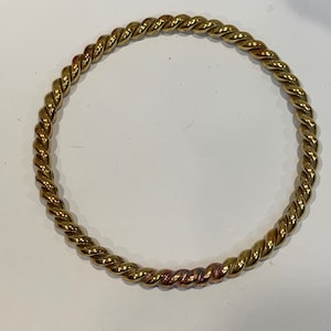 Könnte beinhalten: Ein goldfarbenes, gedrehtes Metallarmband. Das Armband hat ein seilartiges Design, wobei die Metallstränge eng miteinander verdreht sind. Das kreisförmige Armband ist ein schlichtes, aber elegantes Schmuckstück.