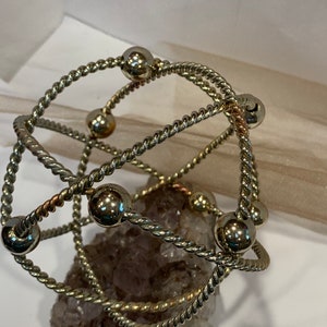 May include: A silver and gold wire bracelet with multiple strands and round beads. The bracelet is resting on a purple and white crystal.