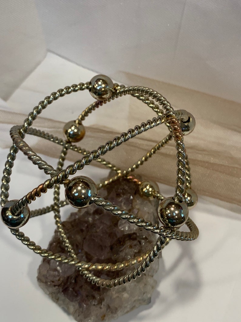 May include: A silver and gold wire bracelet with multiple strands and round beads. The bracelet is resting on a purple and white crystal.