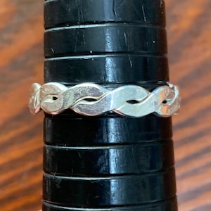 May include: A silver-colored ring with a braided design. The ring is displayed on a black ring sizer. The braided pattern is made up of interlocking, curved metal segments, creating a continuous loop. The ring's surface reflects light, highlighting its metallic sheen.