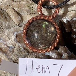 May include: A copper wire wrapped pendant with a round, iridescent, gray stone. The pendant is hanging from a black cord. The text "Item 7" is written on a white piece of paper.