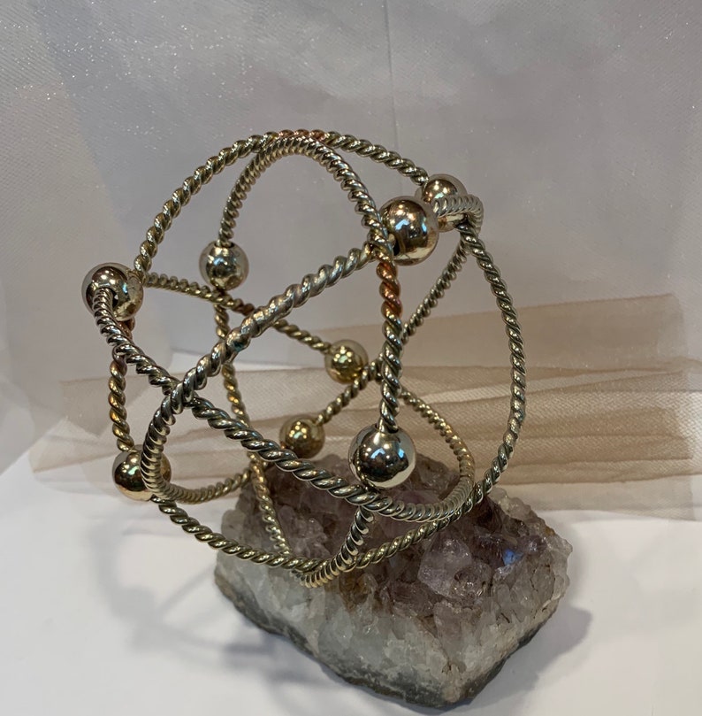 May include: A silver and gold metal sculpture with a geometric design. The sculpture is made of twisted wire and has small spheres at the ends of the wire. The sculpture is sitting on a large, clear quartz crystal.