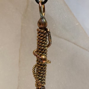 May include: A gold-toned pendant necklace with a long, twisted wire design. The pendant is made of multiple strands of wire that are twisted together to create a rope-like effect. The pendant is attached to a black cord.