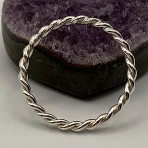 May include: A thin, circular ring crafted from polished silver metal with a continuous twisted rope texture. The band features a uniform, metallic sheen and a consistent spiral pattern throughout its entire circumference, creating a simple, reflective design.