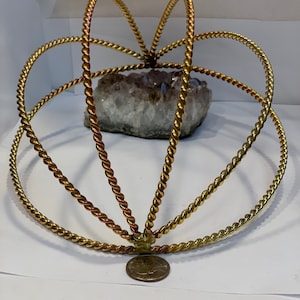 Puede incluir: Una esfera decorativa de metal dorado con un diseño de cuerda trenzada. La esfera está compuesta por anillos interconectados y soportes verticales. Un gran cristal gris y blanco es visible en el interior. Una moneda está en la base.