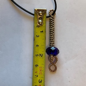 May include: A handmade necklace with a dark cord, featuring a gold-toned metal pendant. The pendant has a coiled design, a blue faceted bead, and a cylindrical section. The necklace is approximately 3 inches long.