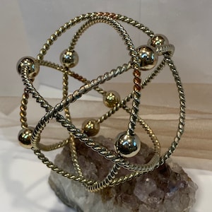 May include: A gold-toned metal bracelet with a twisted rope design and round beads. The bracelet is displayed on a piece of amethyst.