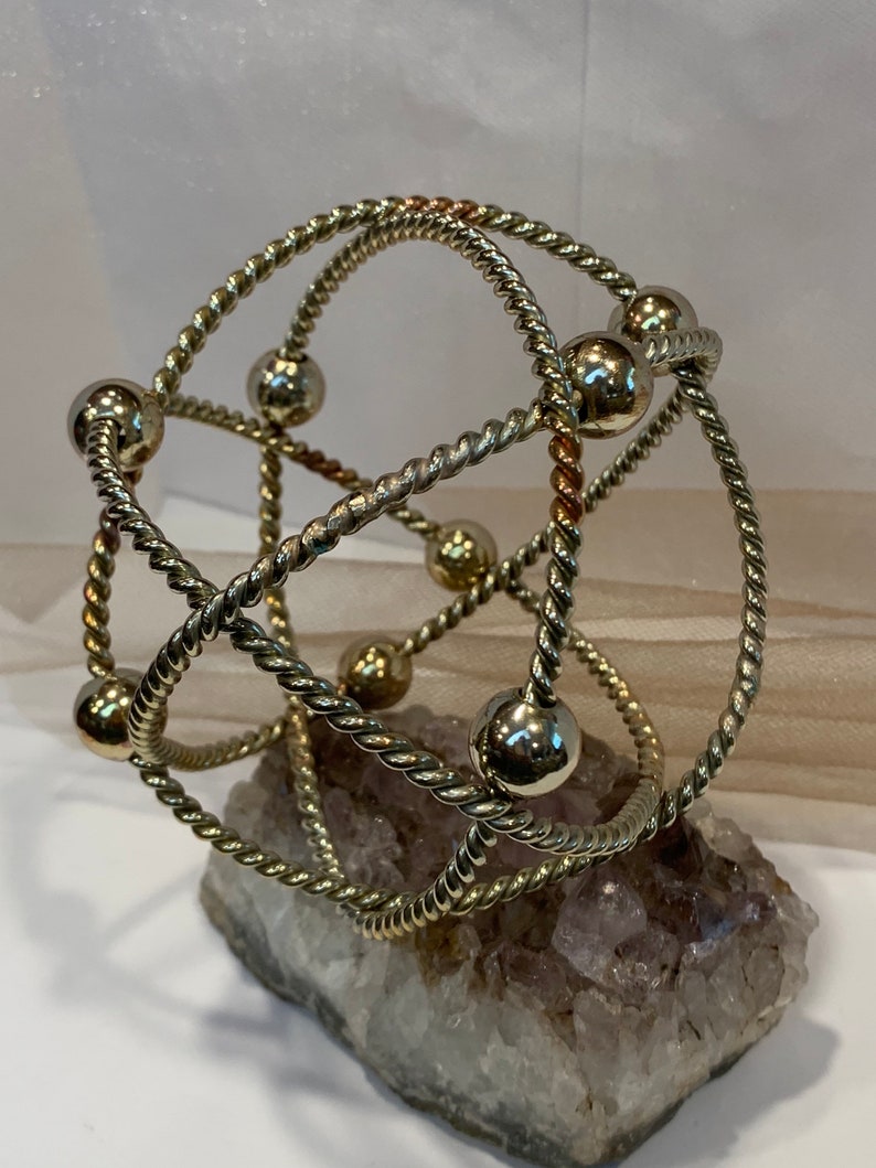 May include: A gold-toned metal bracelet with a twisted rope design and round beads. The bracelet is displayed on a piece of amethyst.
