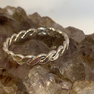 May include: A silver ring featuring a continuous twisted rope design. The band is crafted from a polished metallic material with a reflective surface, showcasing a repeating braided pattern that encircles the entire circumference of the ring.