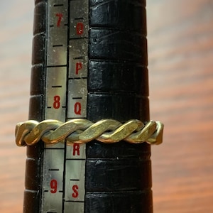 May include: A metallic ring with a twisted rope design positioned on a black ring mandrel. The ring rests between the size 8 and 9 markings on the metal scale, which features red numbers and letters indicating specific ring sizes for measurement.