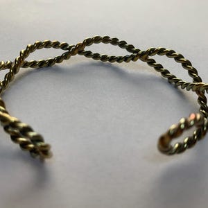 May include: A gold-toned metal cuff bracelet featuring a twisted rope design. The bracelet is constructed from two intertwined strands of metallic wire that form an open, repeating pattern, creating a continuous braided effect around the wrist.