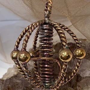 May include: A gold-toned metal pendant with a coiled spring and multiple spheres. The pendant is suspended from a twisted wire loop.