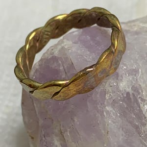 May include: A gold-colored ring with a braided design. The ring is made of metal and has a textured surface. The ring is set against a light purple background.
