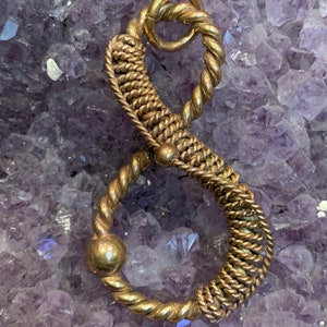 May include: A gold-colored metal pendant shaped like the number eight, with a twisted rope design and two round beads. The pendant is photographed on a bed of purple amethyst crystals.